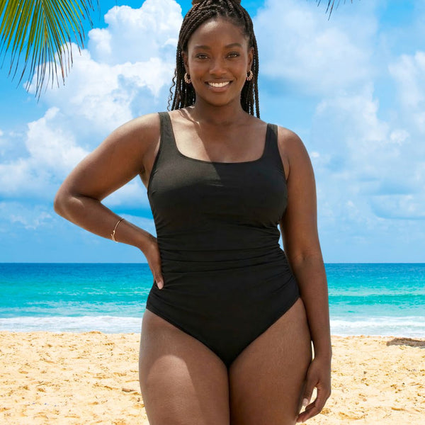 Woman in a black swimsuit standing on a beach with blue sky and ocean in the background